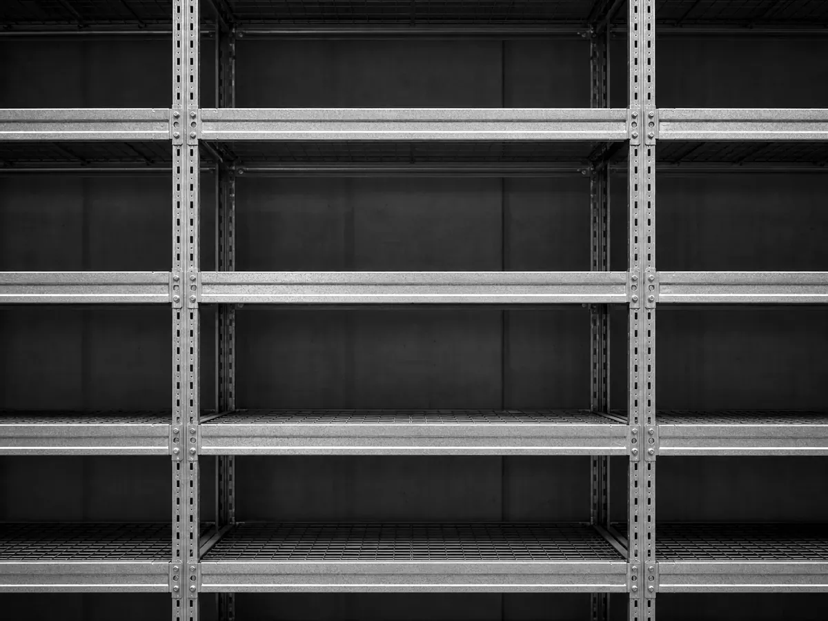 Eine frontale Detailaufnahme eines galvanisierten Stahlregalsystems im Lager — vertikale Pfosten und horizontale Träger bilden ein gleichmässiges Raster, die Stahloberfläche zeigt feine Werkstattstruktur.
