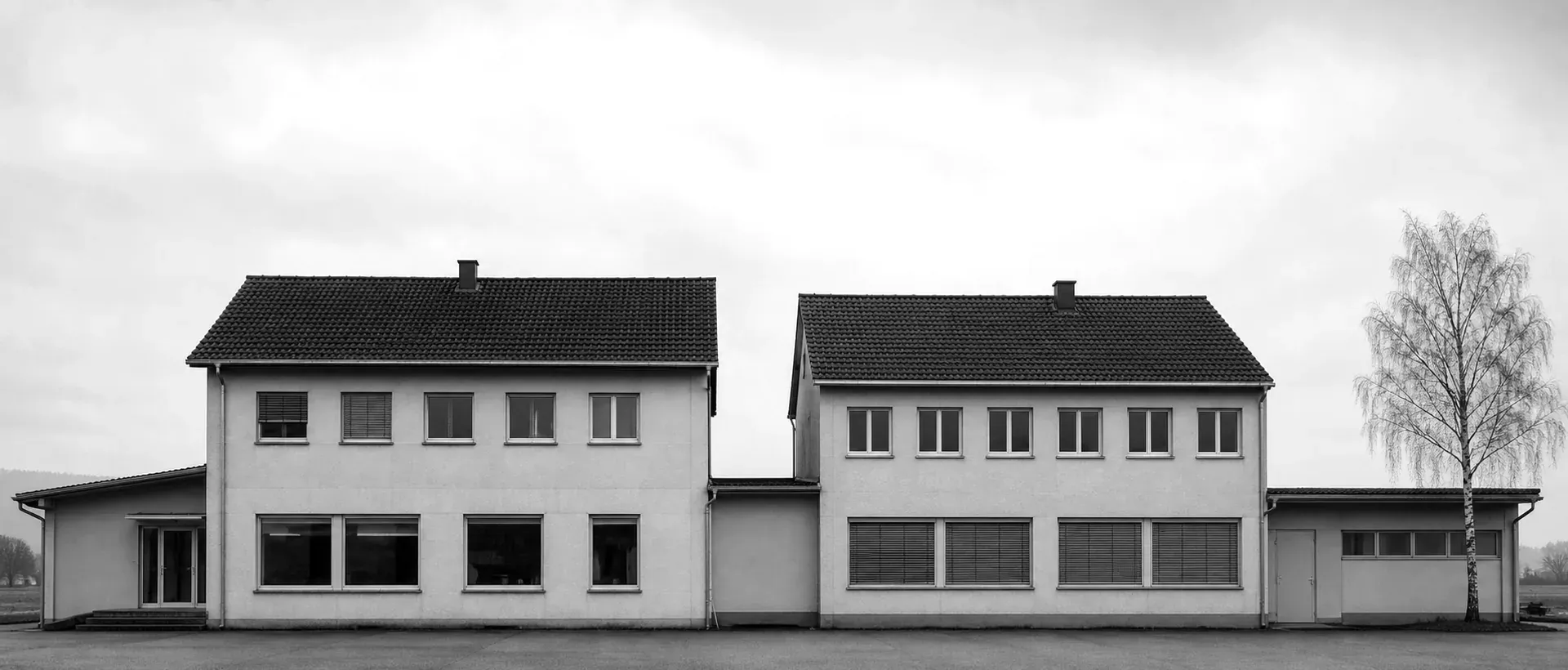 Eine frontale typologische Aufnahme zweier nebeneinanderstehender schlichter Schweizer Geschäftsgebäude im flachen Mittelland-Tageslicht: gerade verputzte Fassaden, gleichmässig verteilte Fenster, leerer grauer Himmel darüber.