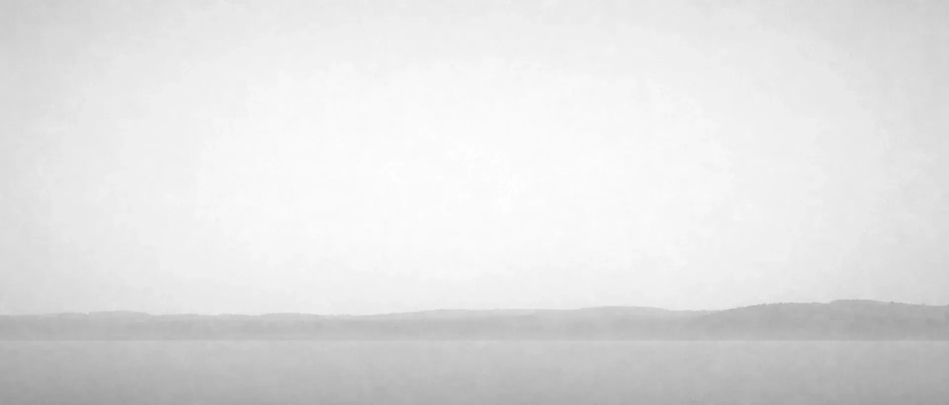 Eine fast abstrakte ruhige Horizontstudie im Seeland-Morgennebel: ein flacher Streifen weicher Dunst unten, weiter Verlauf hellgrauer Himmel darüber.