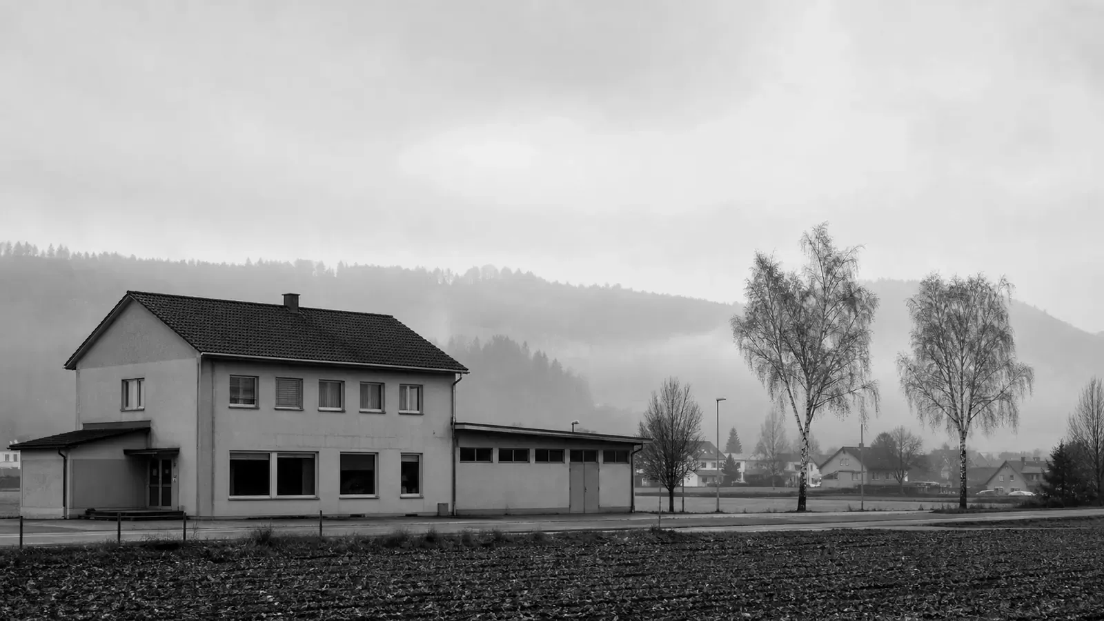 Ein schlichtes zweistöckiges Geschäftsgebäude in einem kleinen Seeland-Dorf am frühen Morgen, leichter Nebel über flachem Ackerland, sanft bewaldete Jura-Hügel in der Ferne — ländliches Kanton Bern.